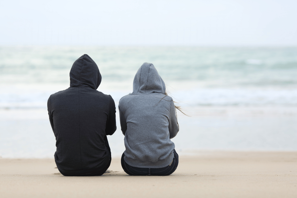 The backs of two friends in sweaters sitting on the beach looking at the sea