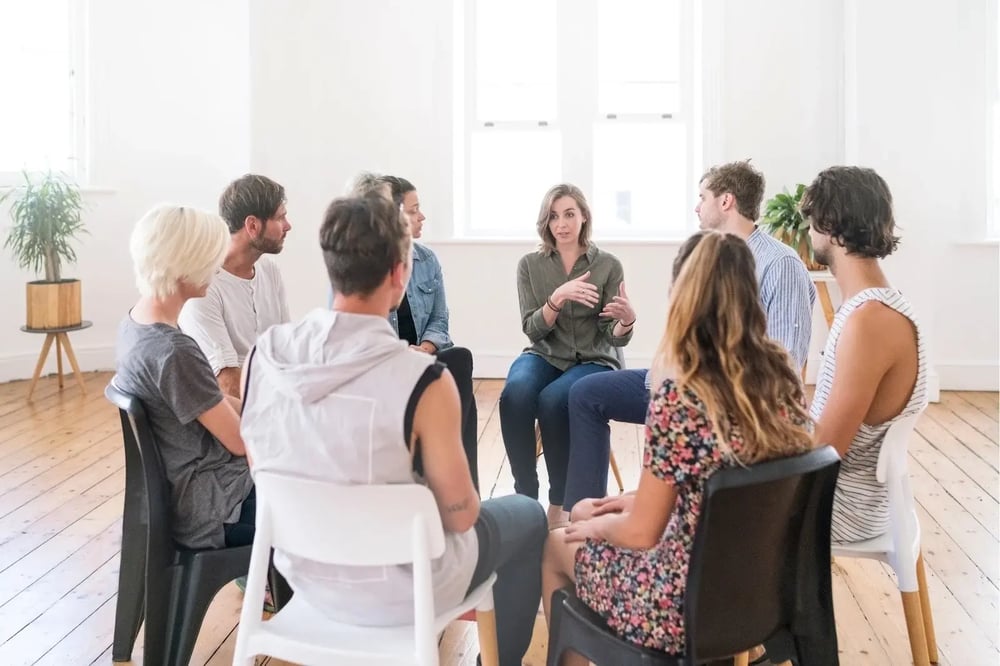 A group therapy session for addiction is underway, with participants listening to a facilitator.