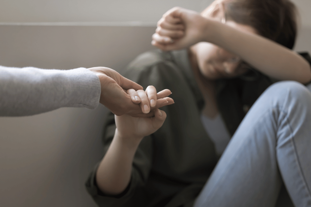 Woman holds her depressed friend’s hand, as the friend sits on the floor, head in her knees.