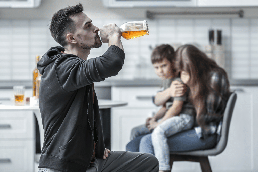 A mother hugs her young son tight as her husband drinks whiskey in front of them