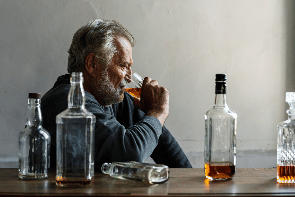 Middle-aged man drinking whiskey from a glass, with several bottles empty on table beside him