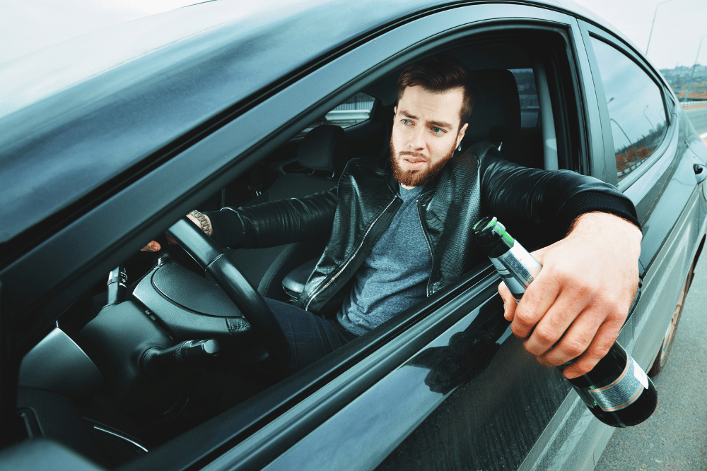 Drunk driver with hand out of his car window as he drives, holding a beer bottle