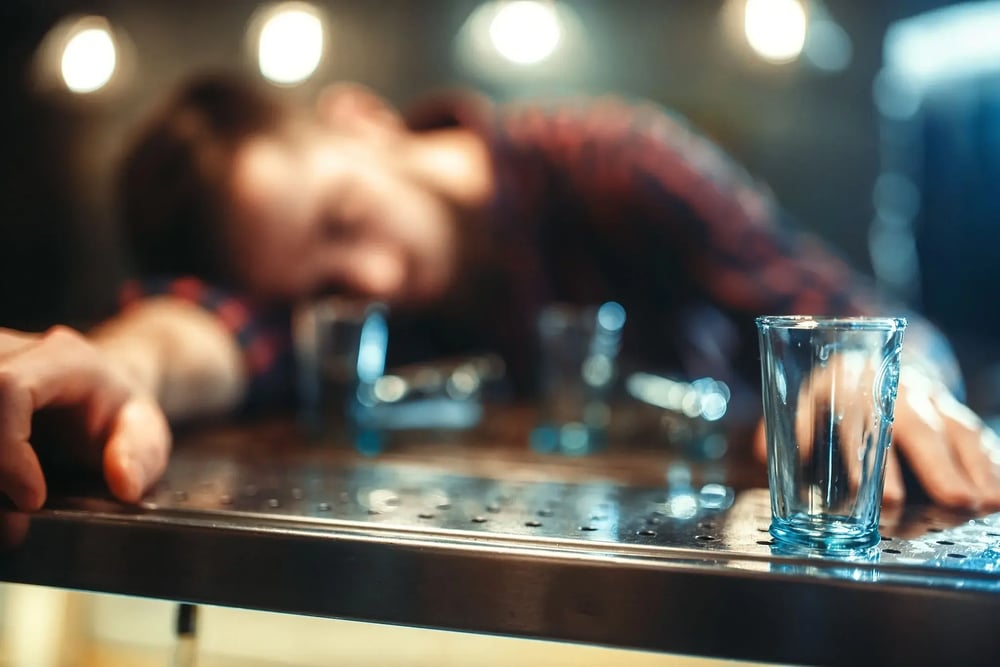Man lying over a bar counter with empty shot glasses around him.