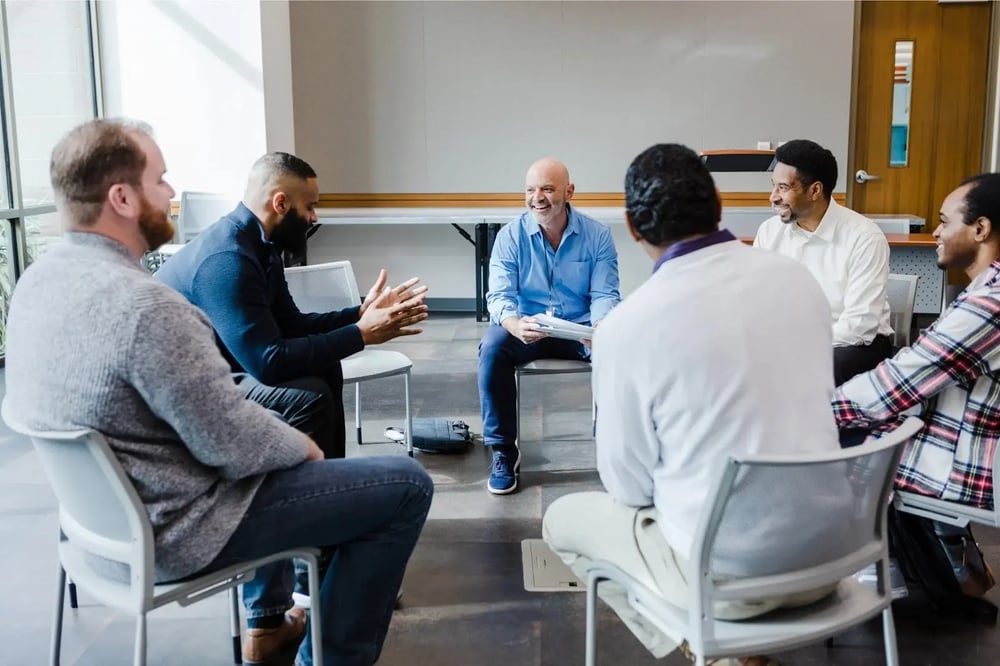 Men sit in a circle listening as one speaks during group therapy for addiction.