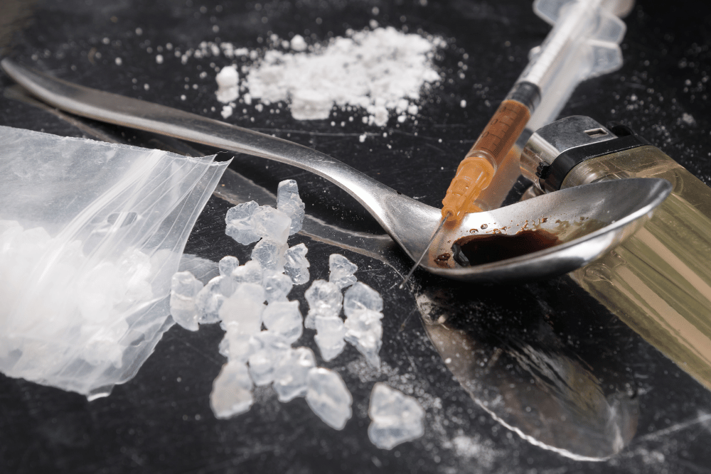Meth with a syringe, spoon, lighter, and plastic bag and white crystals on a dark surface.