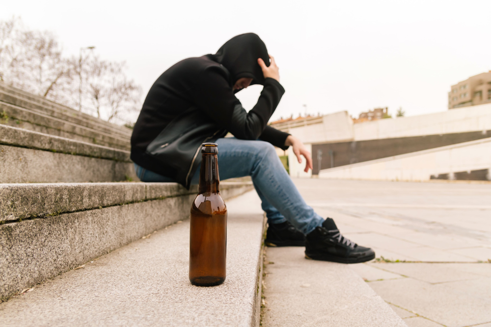 Teenage boy sitting alone on outdoor steps, beside a beer bottle. Relapse may begin with isolation