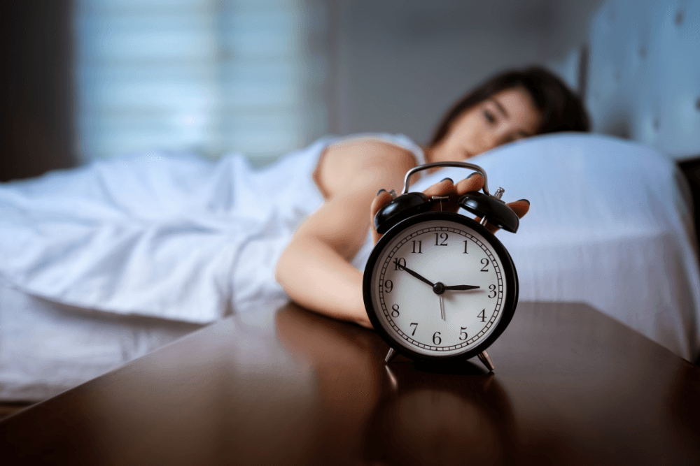 A woman awake in bed, an alarm clock showing 3 AM, seemingly suffering from insomnia