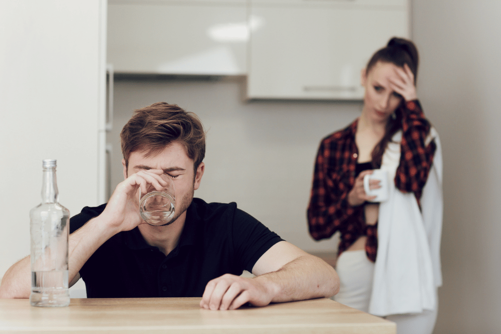 Man drinks vodka by the table, as his partner watches, frustrated