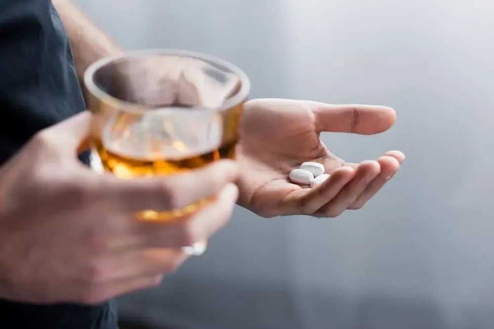 close-up of person's hands holding white pills in one hand and glass of alcohol in second.
